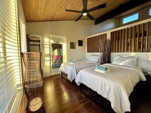 Ein Hotelzimmer mit zwei Betten, einem Deckenventilator, Holzboden und einem großen Fenster mit Jalousien.