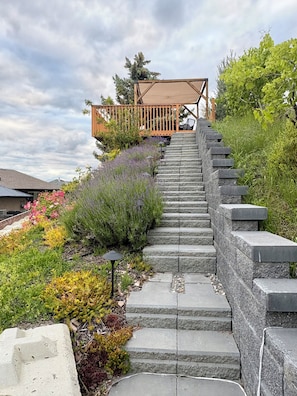 Eine Treppe führt zu einer Holzterrasse mit einer Pergola, umgeben von verschiedenen Pflanzen und Blumen.