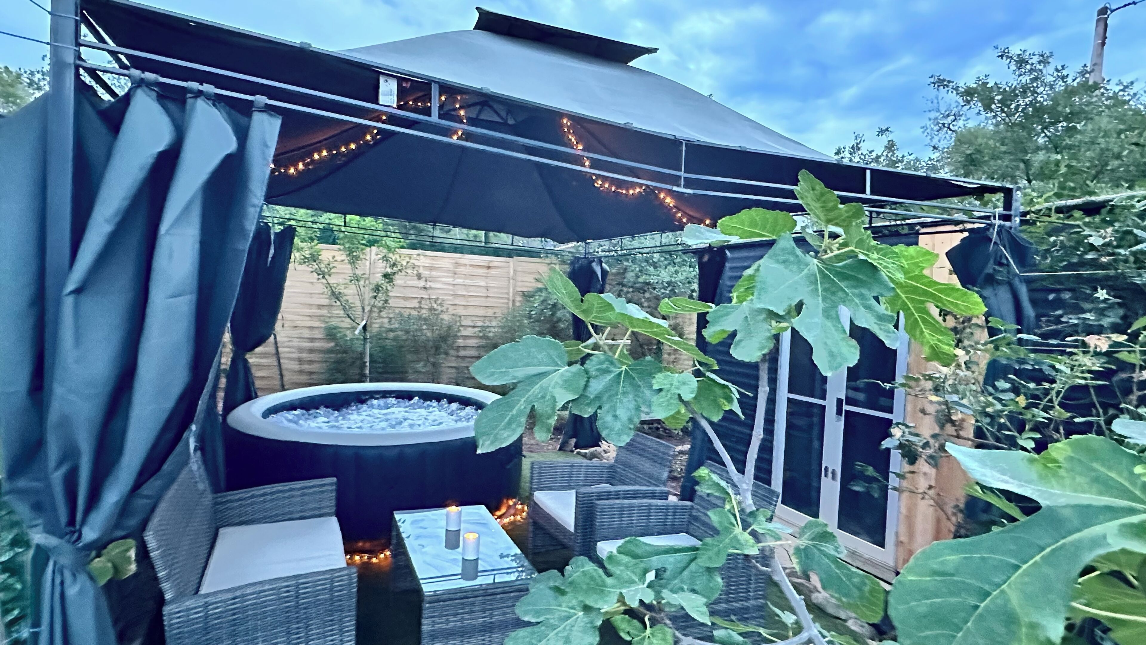 En udendørs terrasse med pejse, fletmøbler og et overdækket område med lyskæder.