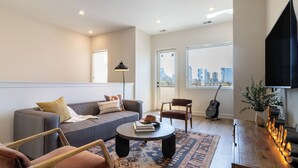 Ein Wohnzimmer mit einer Couch, Sesseln, einem Couchtisch, einer Gitarre, einem Fernseher und Blick auf die Stadt.
