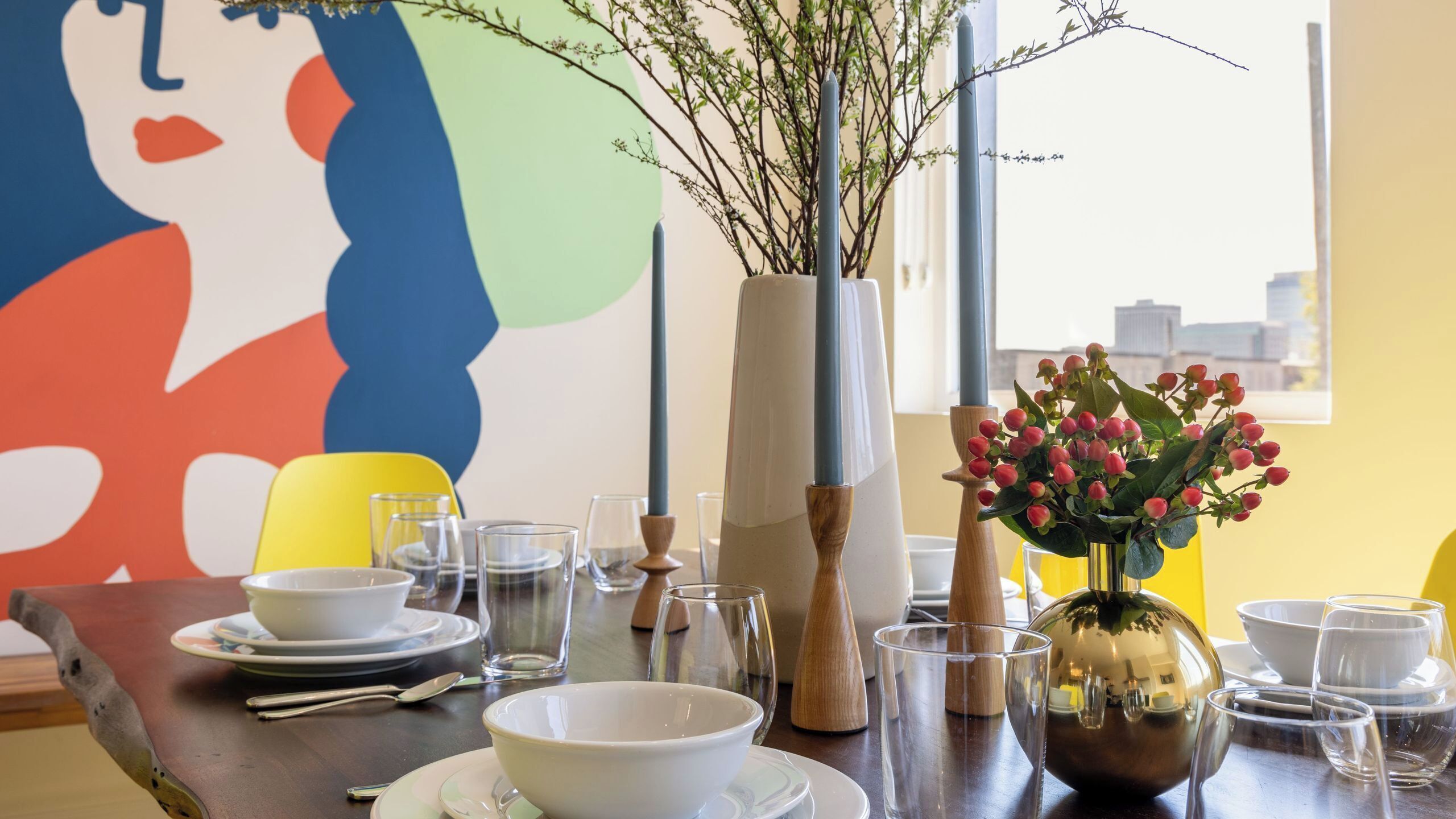 Een eettafel gedekt met witte serviesgoed, glazen en een vaas met rode bloemen.