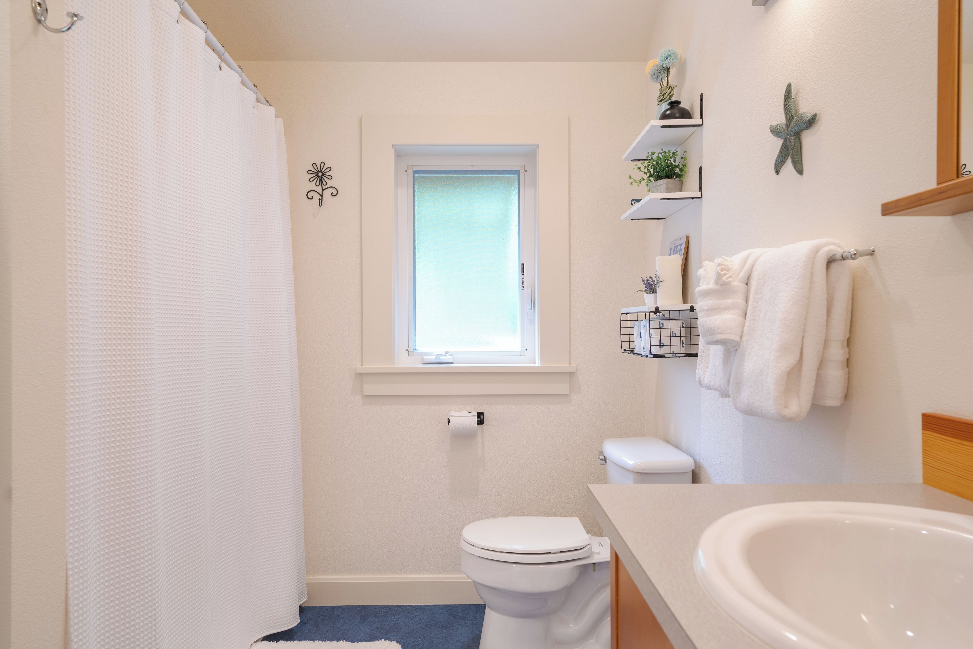 Une salle de bain avec un rideau de douche, des toilettes, un lavabo, une fenêtre et des objets décoratifs placés sur des étagères.