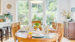 A dining area with a round wooden table set for breakfast, surrounded by chairs, with a view of greenery through large windows. - Walk to Beach + Relax in Hot Tub + Dine on the Deck Loli's Place by Avantstay (Arch Cape)