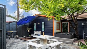 A patio area with stone flooring, a table with benches, a hot tub, and a covered seating area with a blue umbrella. - Red River by Avantstay Shared Pool + Hot Tub 20 Minutes to Downtown Austin (Austin)