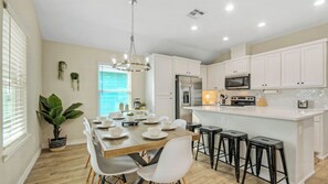 A modern kitchen with white cabinets, a stainless steel refrigerator, a microwave, and a dining area with a wooden table and white chairs.