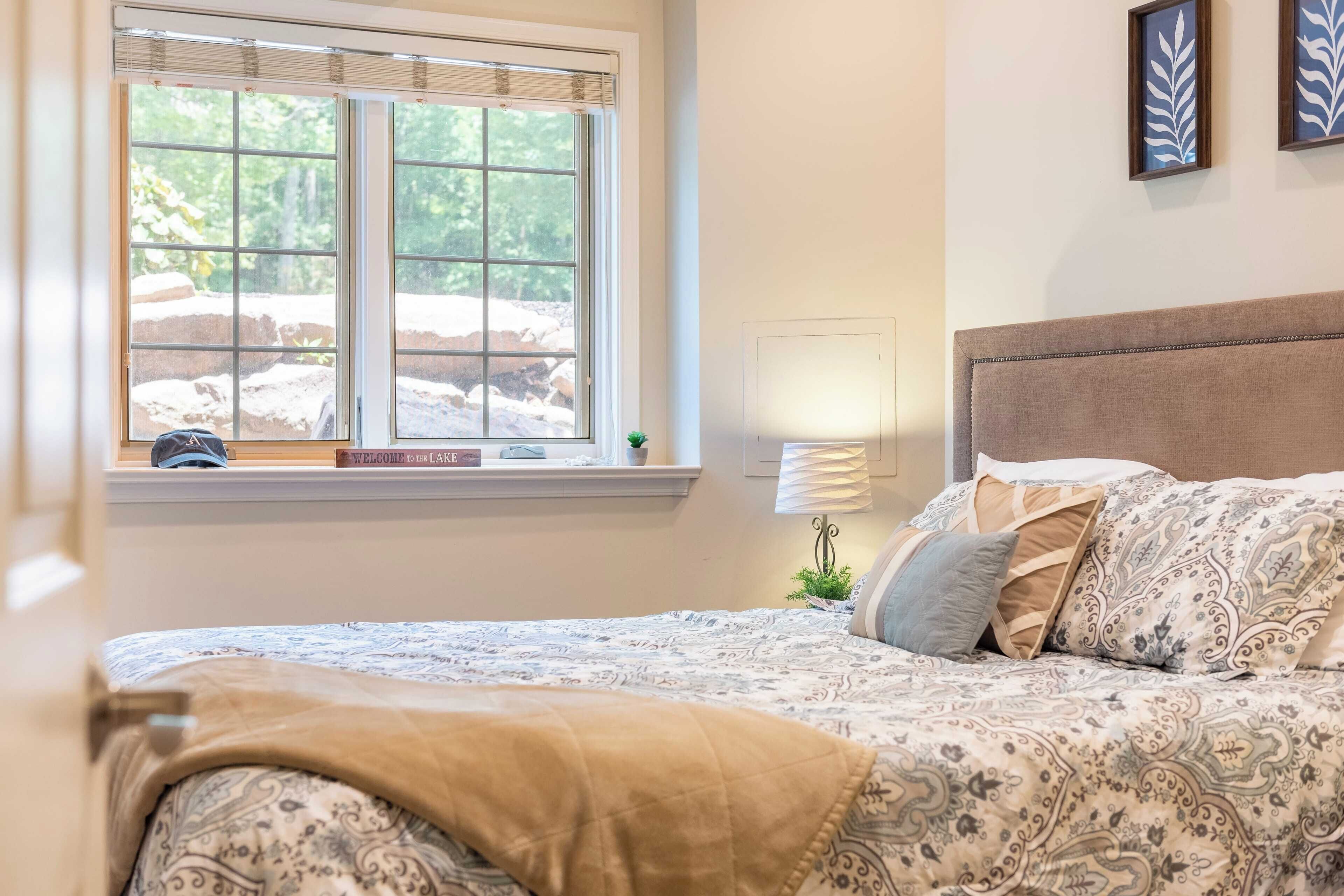 A bedroom with a large bed, a window with blinds, a bedside lamp, and a framed picture on the wall.
