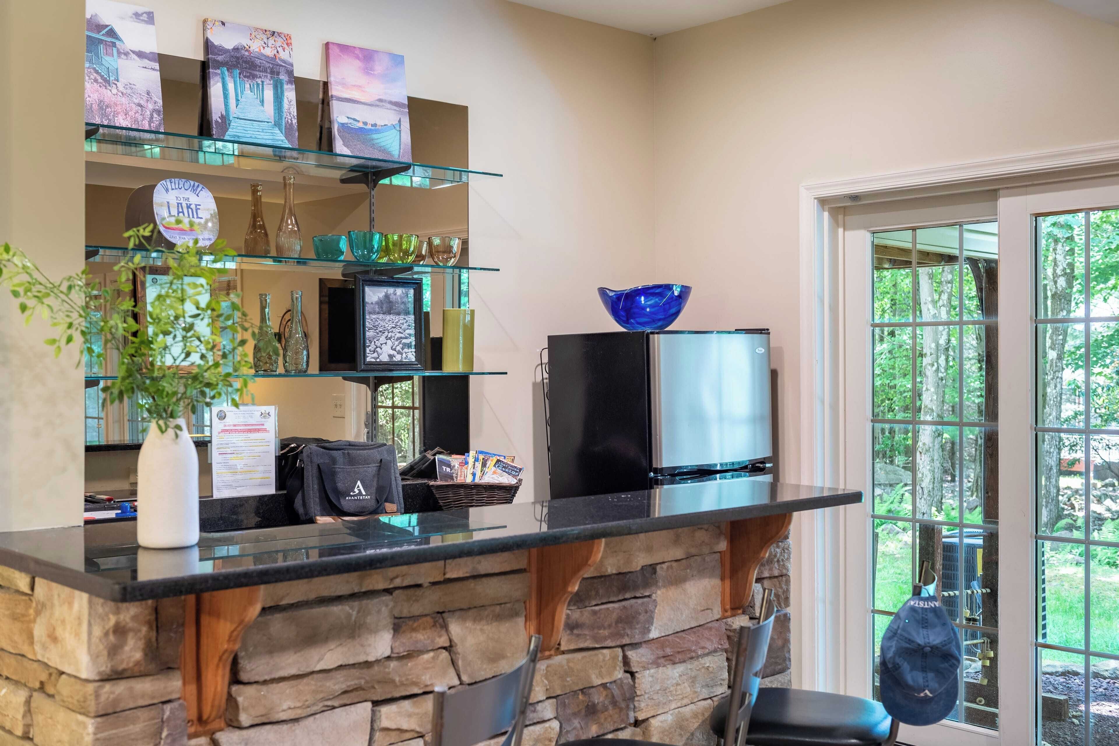 A reception area with a stone counter, glass shelves with decorative items, a black refrigerator, and a view of greenery through large windows.