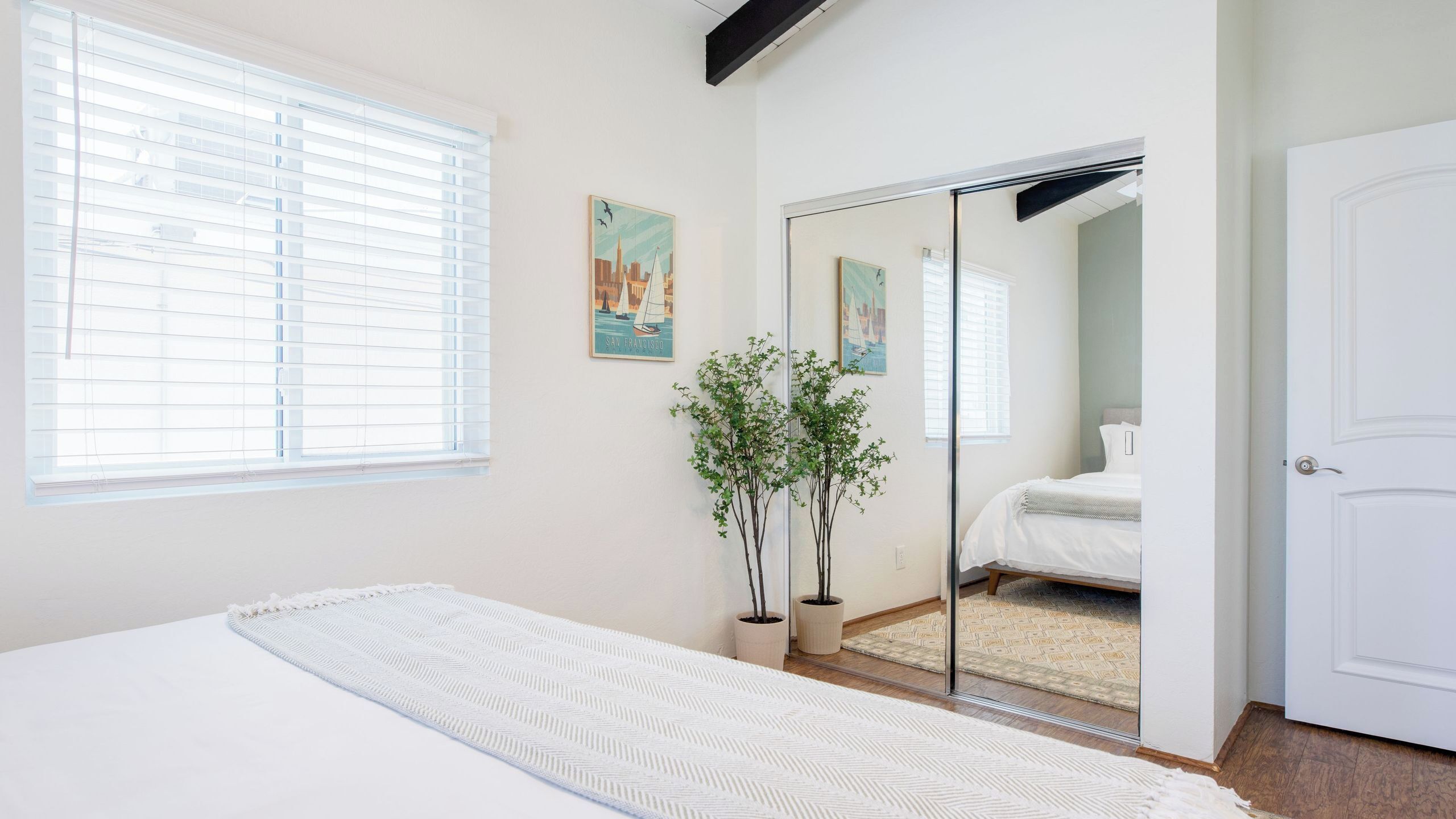 Quarto com cama, espelho, janela com persianas e duas plantas em vasos.