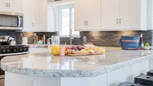 A modern kitchen with white cabinets, a granite countertop, a built-in microwave, and a breakfast bar with fruit and juice. - Surfrider II by Avantstay Walk to the Beach + Balcony (Newport Beach)