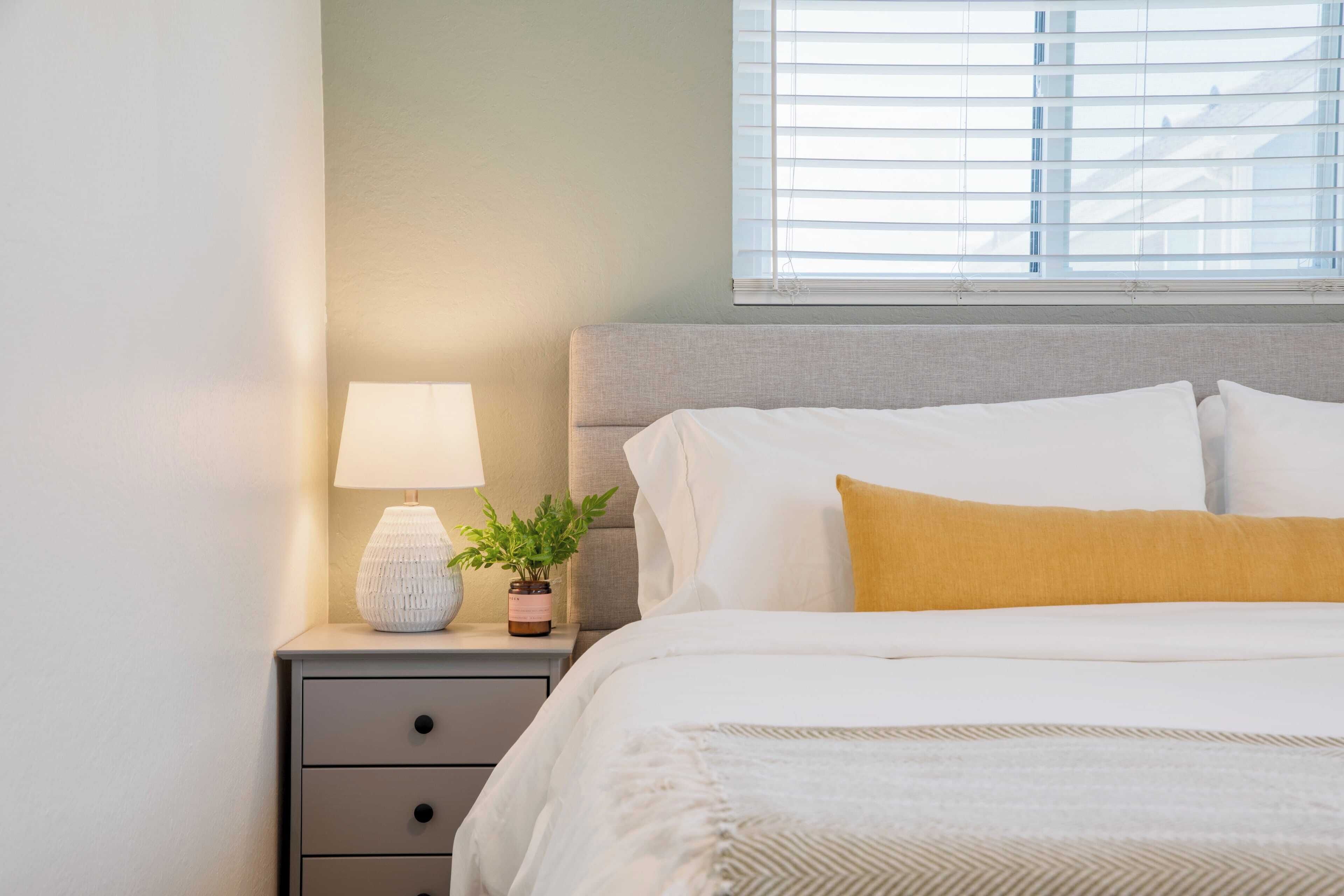 A neatly made bed with white and yellow pillows, a bedside lamp, and a small potted plant on a nightstand.