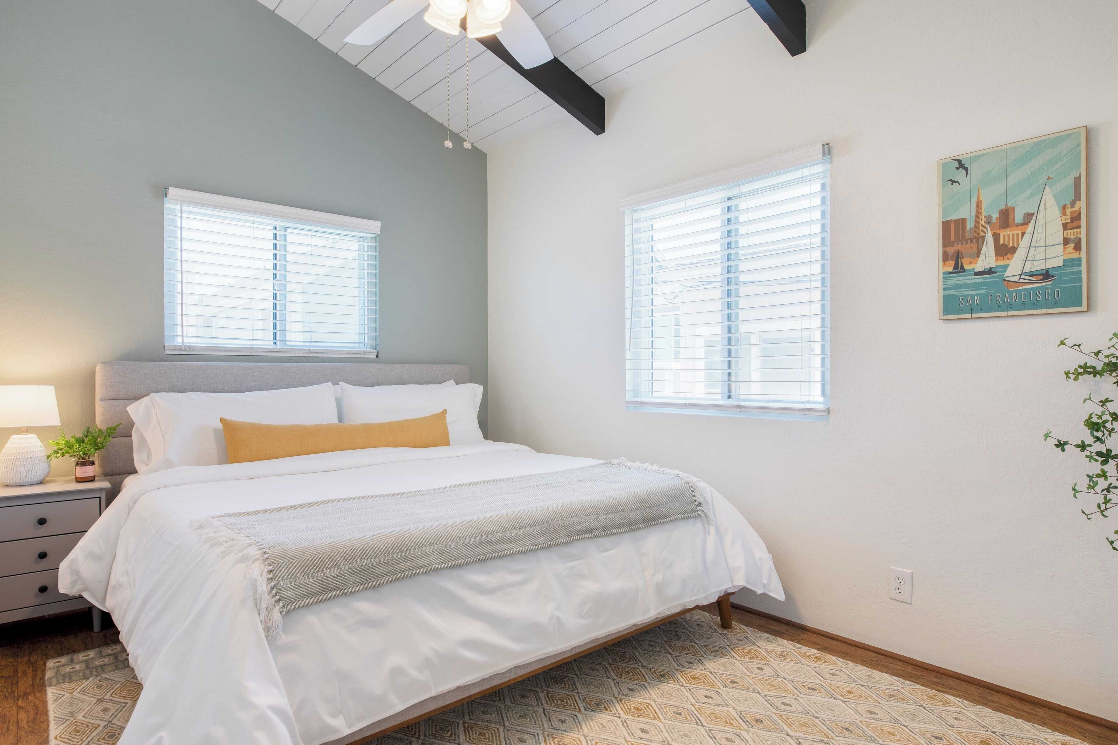 A bedroom with a bed, two windows, a nightstand, a lamp, and a wall art piece.