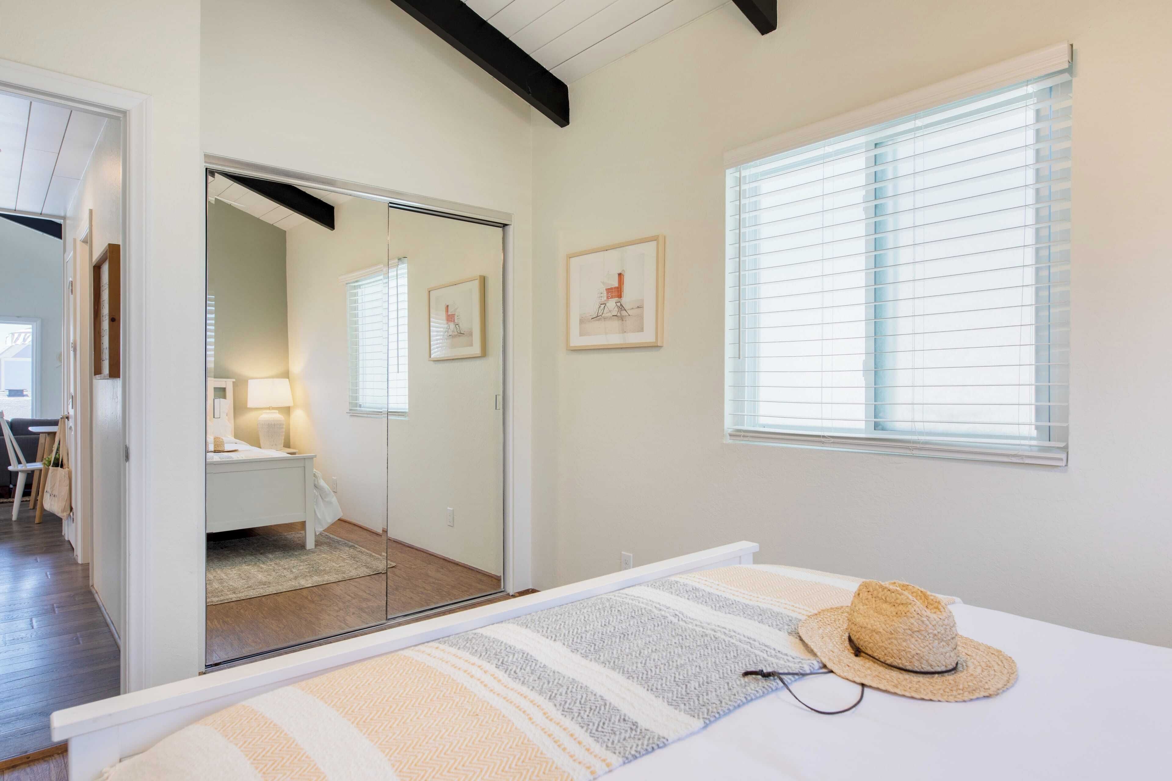 A bedroom with a bed, a mirror, a window with blinds, and a door leading to a bathroom.