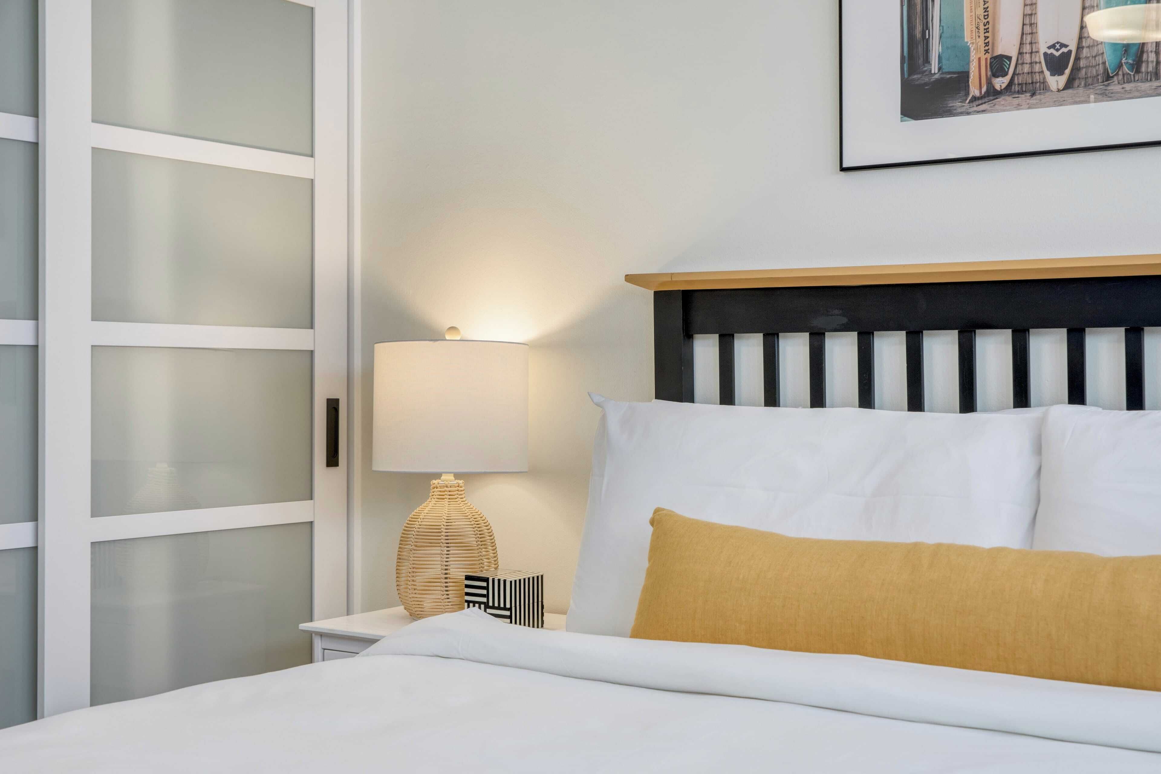 Una cama bien hecha con almohadas blancas y mostaza, una lámpara de noche y un cuadro en la pared.