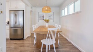 Una cocina moderna con un refrigerador de acero inoxidable, una mesa de comedor de madera con sillas blancas y una lámpara colgante sobre la mesa.