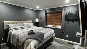 Une chambre à coucher comprenant un lit, une table de chevet, un téléviseur fixé au mur et une fenêtre avec des stores.