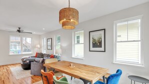 A spacious living room with a wooden dining table, colorful chairs, a ceiling fan, and large windows.