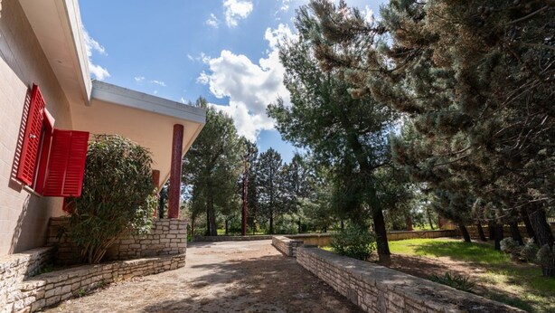 Une cour pavée, un mur en pierre, un bâtiment aux volets rouges et des arbres tout autour.
