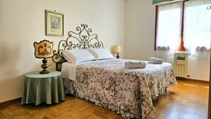 A bedroom with a bed, a bedside table, a lamp, a framed picture on the wall, and a window with curtains. - GuestHost - Spacious and bright 130 sqm apartment, perfect for 4 people, located on the second floor of a building WITHOUT a lift (stairs to access it).The apartment is located 4.1 km from the center of Lucca: in just 13 minutes by bus you can reach the  (Lucca)