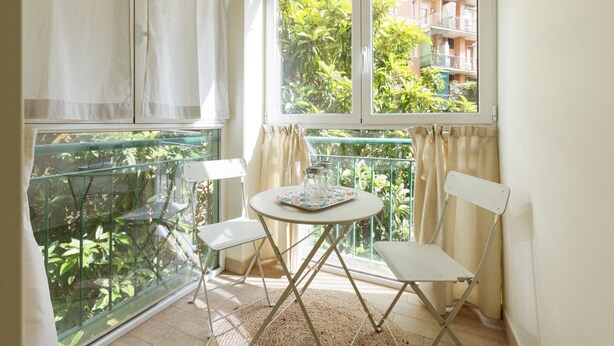 Een balkon met glazen omheining, twee witte stoelen, een klein rond tafeltje, en uitzicht op groen en gebouwen.
