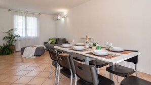 Un comedor con una mesa blanca dispuesta para cuatro personas, rodeada de sillas negras. Un sofá gris y una planta en maceta se ven al fondo.