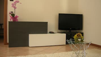 Ein modernes Wohnzimmer mit Flachbildfernseher, einem weißen Sofa, einem Glastisch und einer dekorativen Orchideenpflanze.