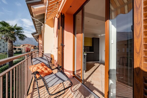 A balcony with a sun lounger, a glass door leading to a kitchen, and a view of mountains.