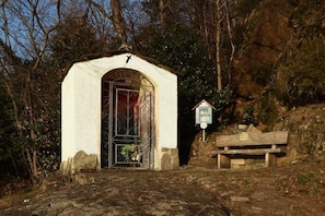 Une petite chapelle blanche, dotée d’une porte en métal et surmontée d’une croix, entourée d’arbres et bordée d’un banc en pierre.