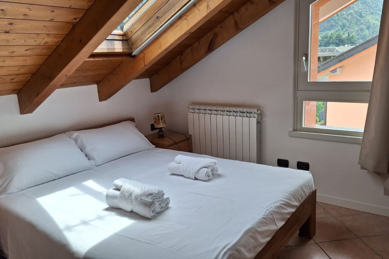 Un dormitorio con techo inclinado, una cama con ropa blanca, un radiador y una ventana con vista a edificios.