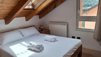 Un dormitorio con techo inclinado, una cama con ropa blanca, un radiador y una ventana con vista a edificios.