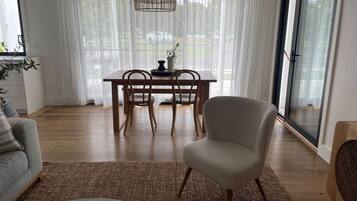 A modern living room with a sofa, a chair, a dining table with chairs, a woven rug, and a large window with sheer curtains.