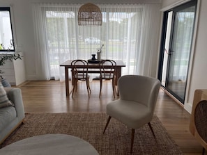 Una sala moderna con un sofá, una silla, una mesa de comedor con sillas, un tapete tejido y un ventanal con cortinas de gasa.