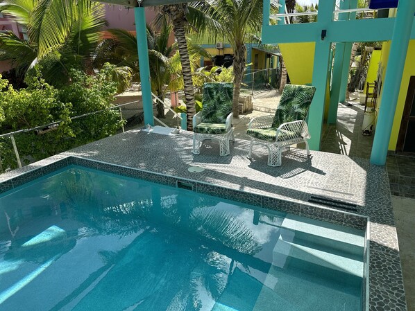 A swimming pool with tiled edges, surrounded by white wicker lounge chairs and palm trees.