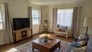 A living room with a TV on a wooden stand, a coffee table, a sofa, a small table with a plant, and a window with curtains. - Cozy 1Bed/1Bath unit, Pets OK, Close to ProMedica, UT. (Toledo)