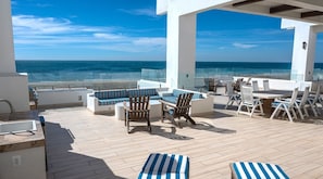 Relax on the ocean-facing terrace with sofas, chairs, and a dining table, surrounded by a glass railing offering breathtaking views of the beach and sea.