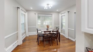 A dining area with a wooden table set for four, surrounded by chairs, with windows and curtains on the walls.
