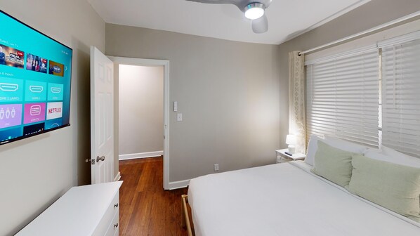 Quarto de hotel com televisão exibindo diversos serviços de streaming, cama com travesseiro verde, mesa de cabeceira com abajur e janela com persianas.