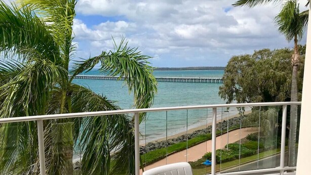 A balcony with a table, two chairs, a bottle of wine, and a glass. The balcony overlooks a beach with palm trees and a pier in the distance. - 180˚ Water View Apartment - Simply Stunning (Urangan)