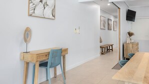 Une pièce moderne et bien éclairée, avec un bureau en bois, une chaise bleue et une longue table à manger. Un tableau encadré orne le mur, et un téléviseur est fixé au-dessus de la table.