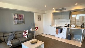 A modern living room with a sofa, coffee table, and a small side table with decorative items. Adjacent is a compact kitchen with white cabinets and a window overlooking a cityscape. - Amazing City Skyline Apartment - Pool, Gym, Sauna. (Adelaide)