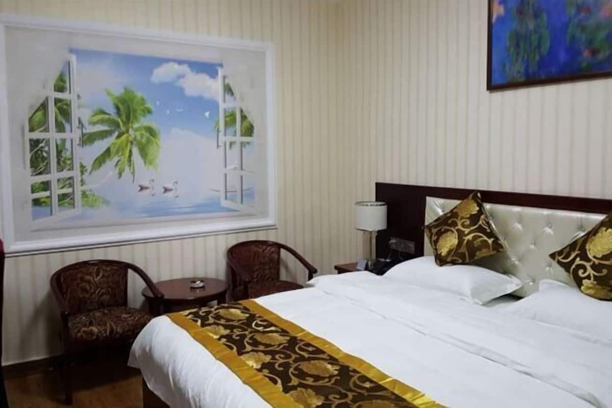 Une chambre d’hôtel avec un lit, deux chaises, une petite table, une fenêtre donnant sur des palmiers et un tableau au mur.