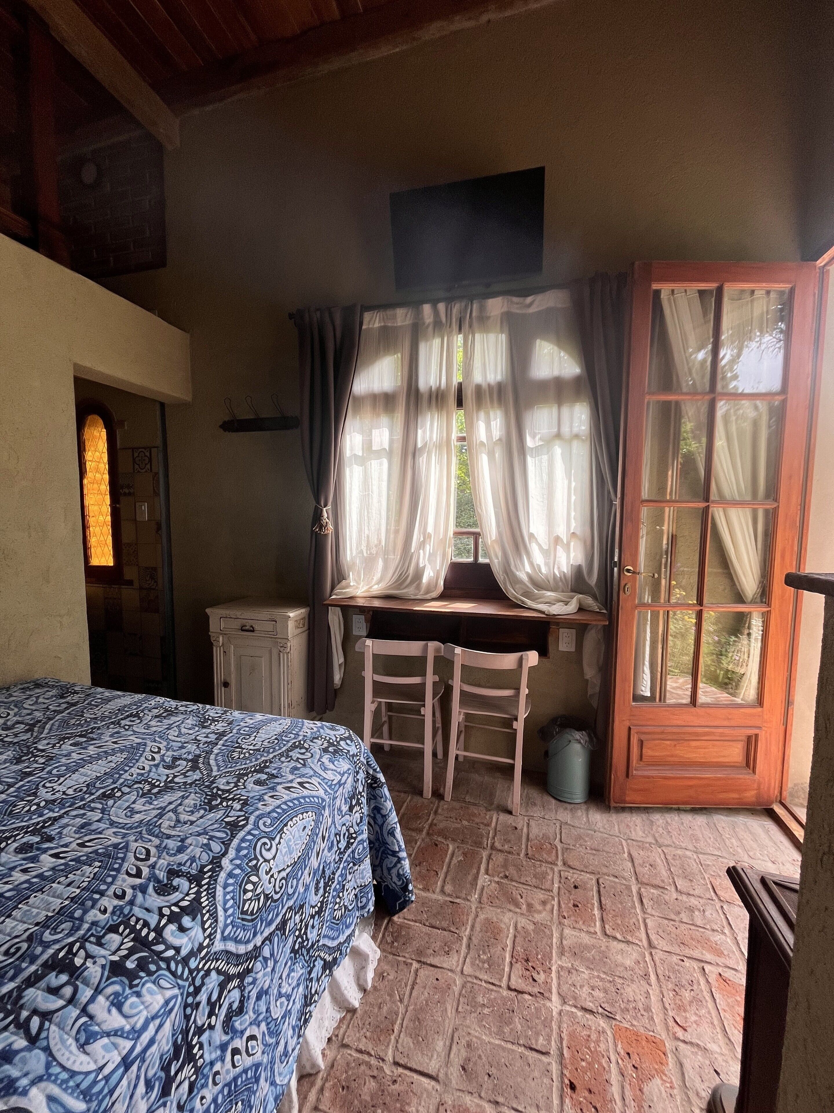 A bedroom with a bed, a desk, two chairs, a window with curtains, and a door.
