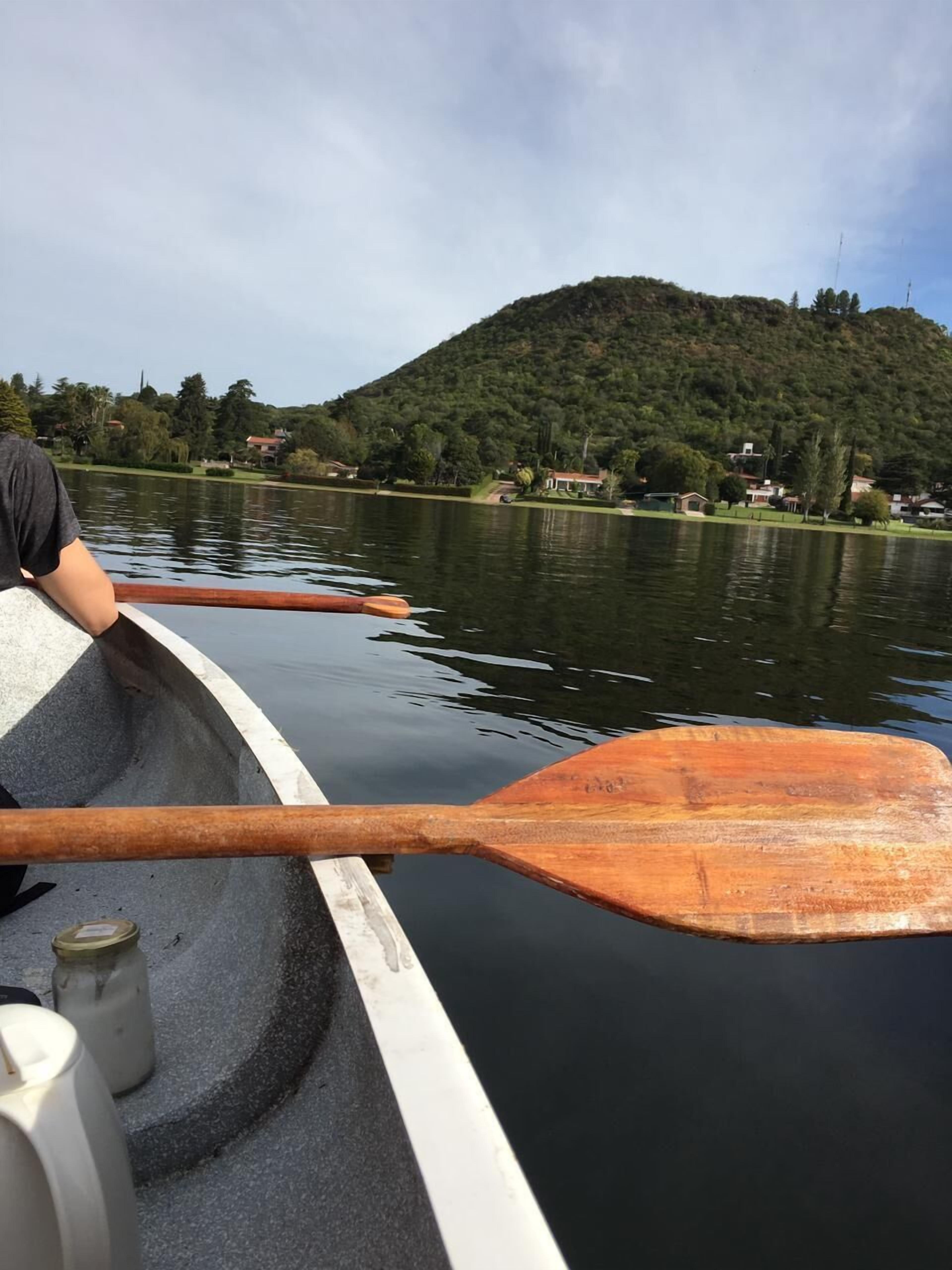 Un bote con una persona apoyada en el borde, remos de madera y un lago tranquilo con una colina al fondo.