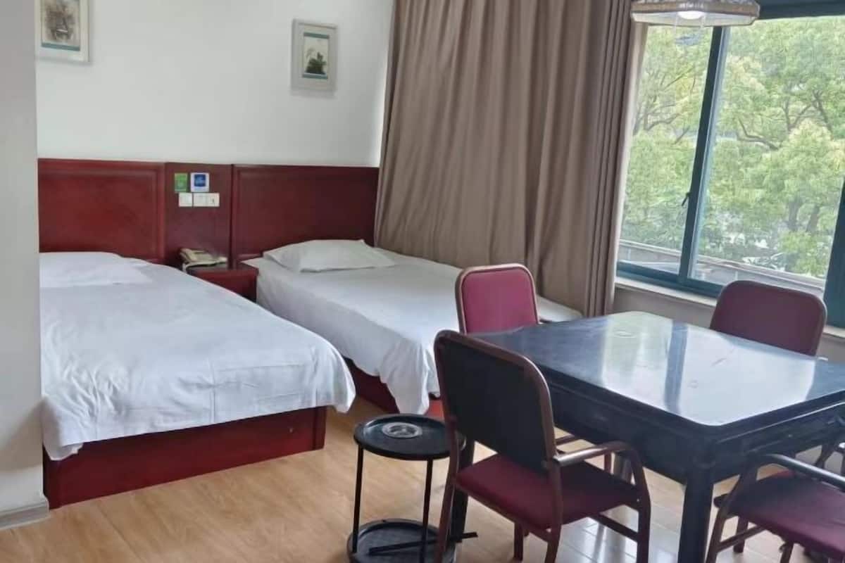 A hotel room with two single beds, a small table with four chairs, a window with a view of trees, and a curtain.
