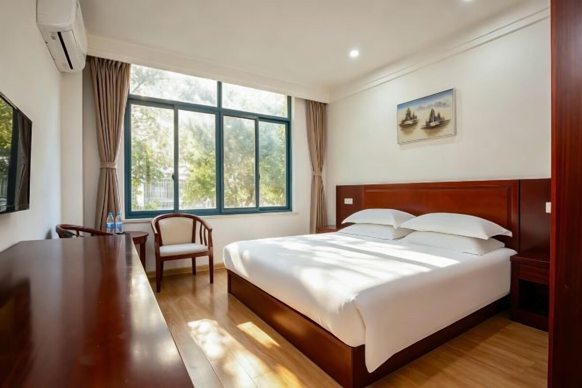 A hotel room with a bed, a desk, a chair, a window with curtains, and a painting on the wall.
