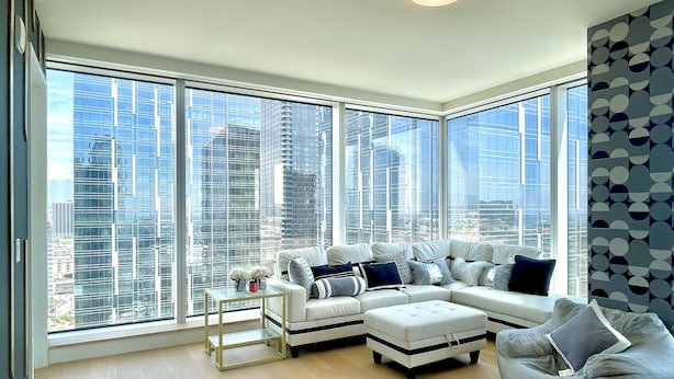 Una sala moderna con un sofá seccional en forma de L, una mesita auxiliar pequeña y un sillón tipo bean bag. La habitación tiene ventanas de piso a techo con vistas a la ciudad.