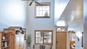 Un salón espacioso con techo alto, vigas de madera, ventilador de techo, un ventanal grande y una escalera que conduce a un nivel superior.