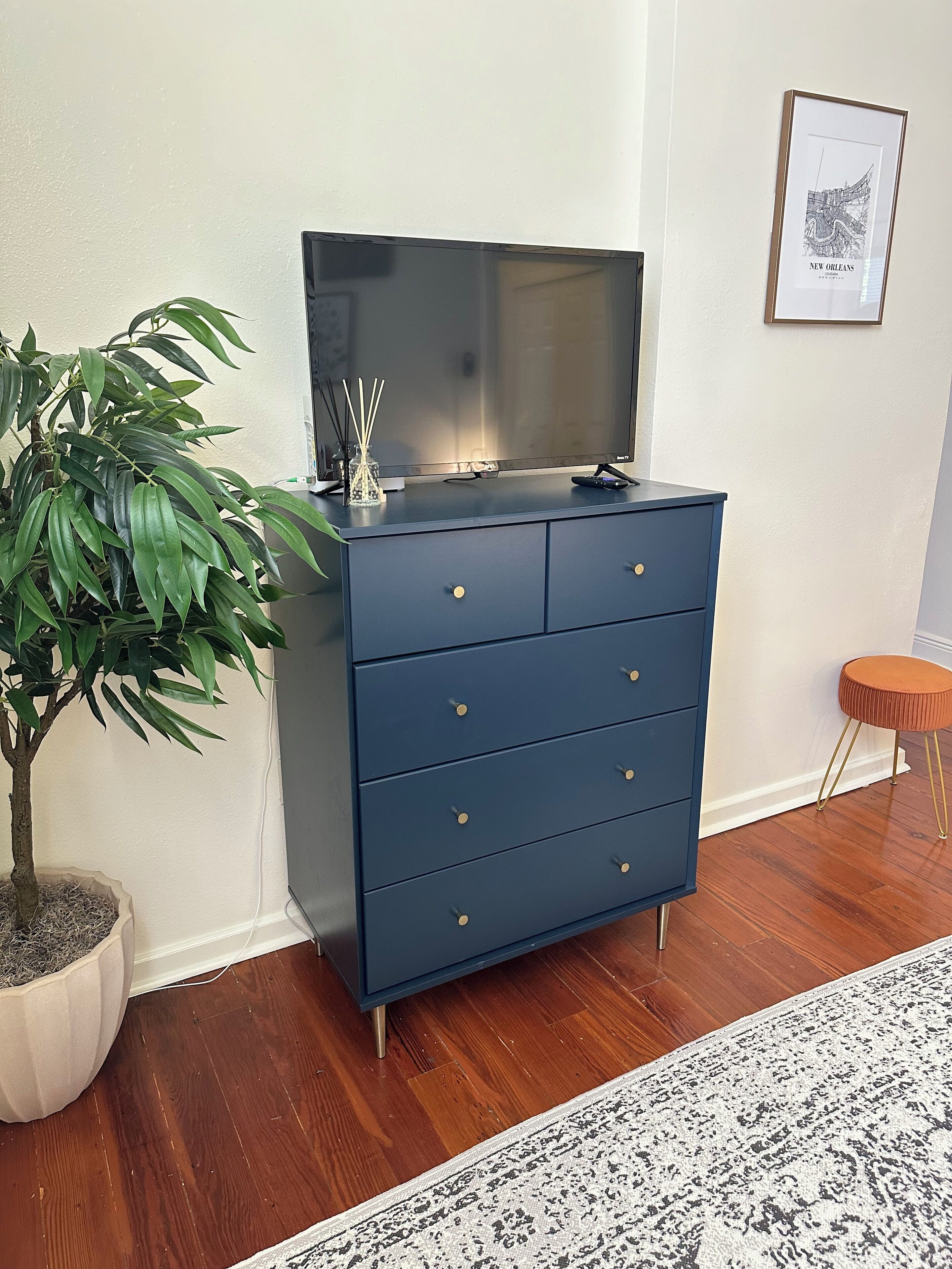 Ein dunkelblauer Kommode mit Schubladen, ein Blumentopf, ein gerahmtes Bild an der Wand und ein kleiner Hocker.