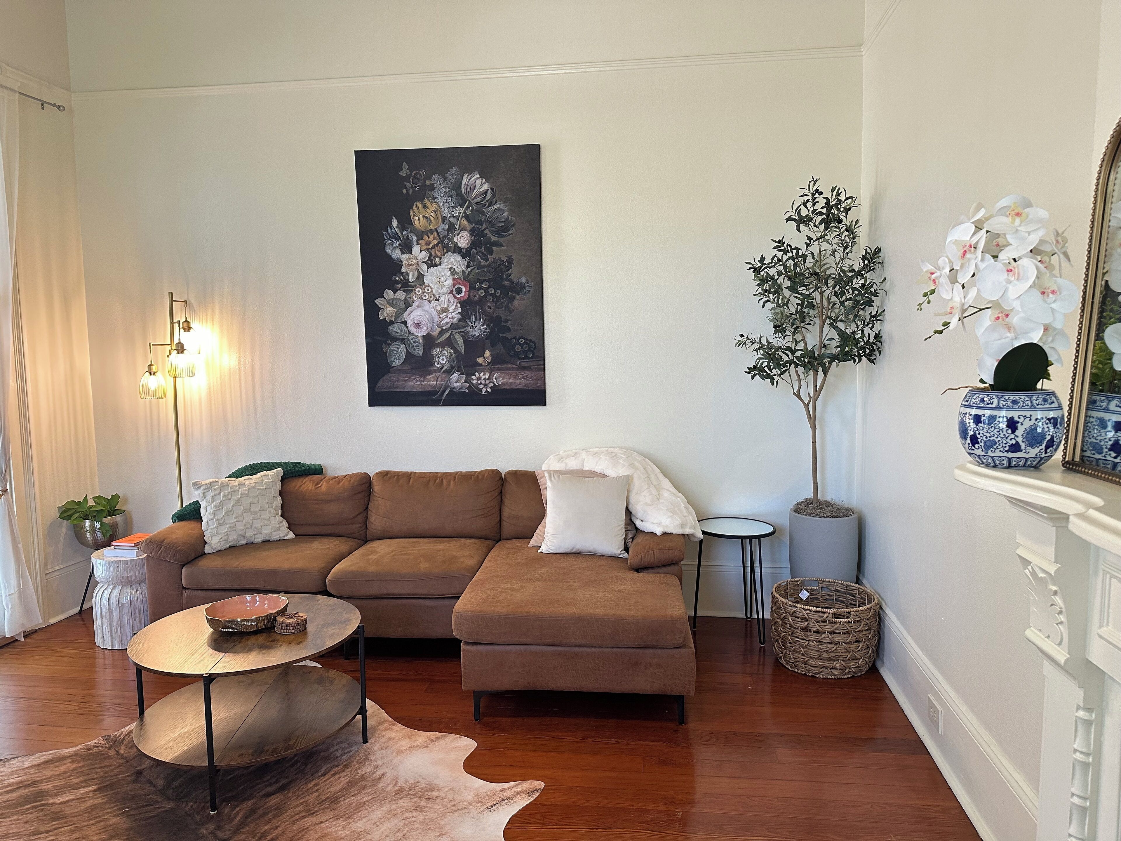 A living room with a brown sectional sofa, a round coffee table, a floor lamp, a painting of flowers, a potted plant, and a mirror on the wall.