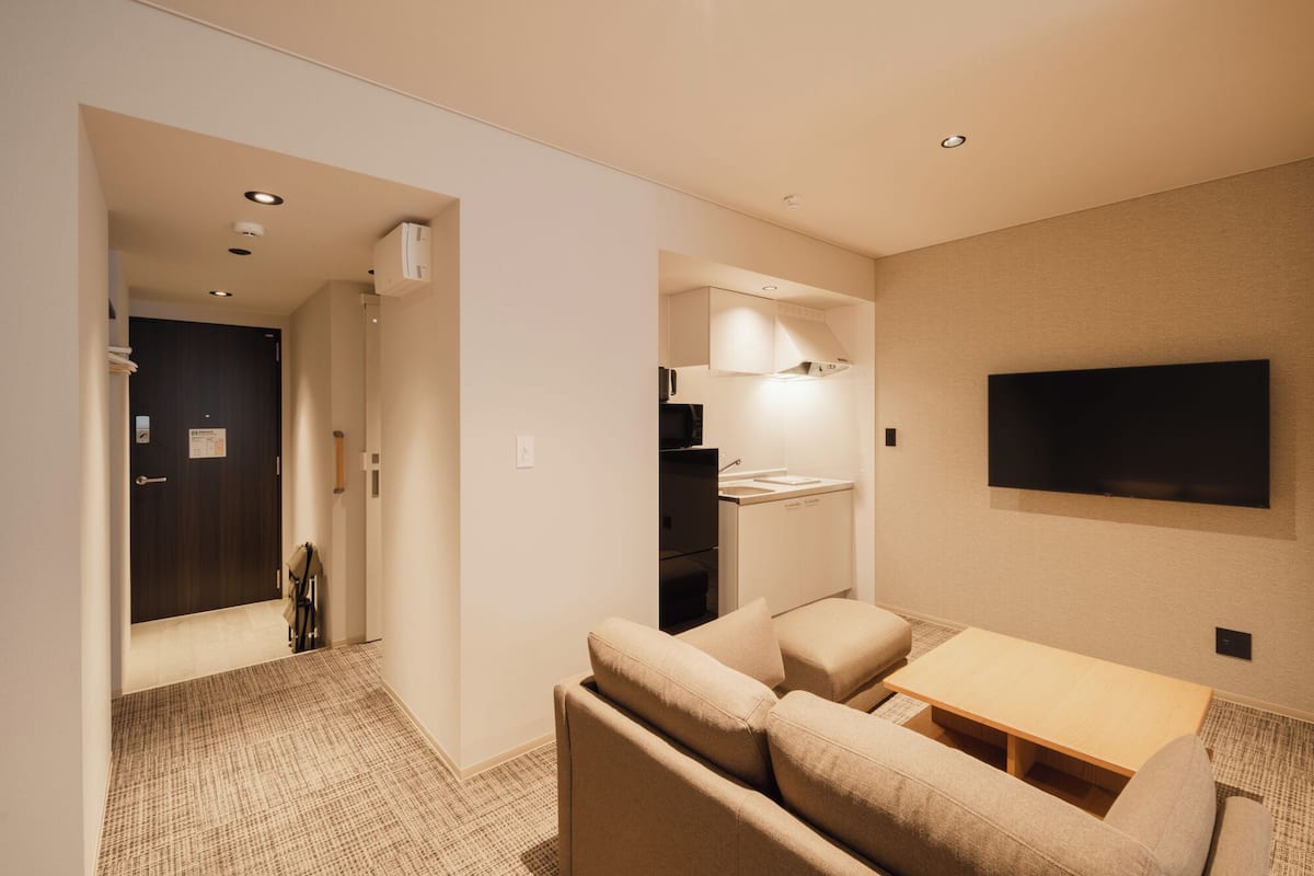 Ein modernes Hotelzimmer mit einer Couch, einem kleinen Tisch, einem an der Wand montierten Fernseher und einer Küchenzeile.