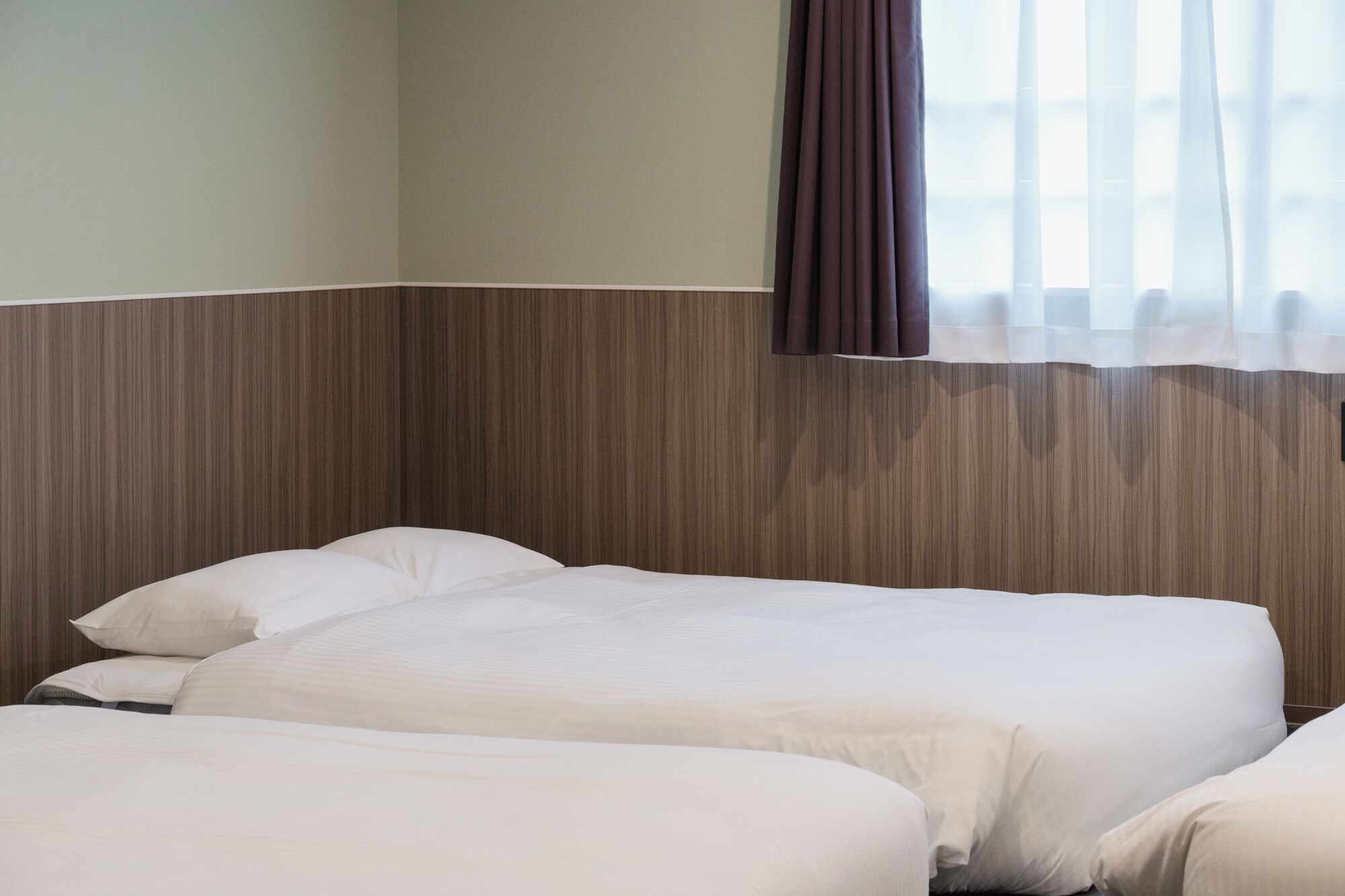 A hotel room with two beds, a wooden headboard, a window with curtains, and a plain wall.